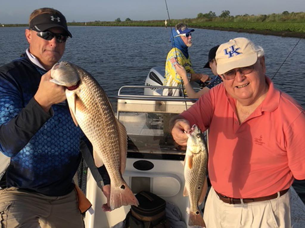 Two men on a boat each holding a Redfish, one’s is much bigger. Two more people are in the background, and one’s fishing.