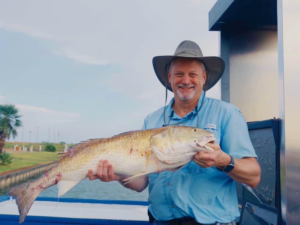 A man in a wide-brimmed hat and blue shirt's standing on a boat, holding a large Redfish and smiling at the camera.