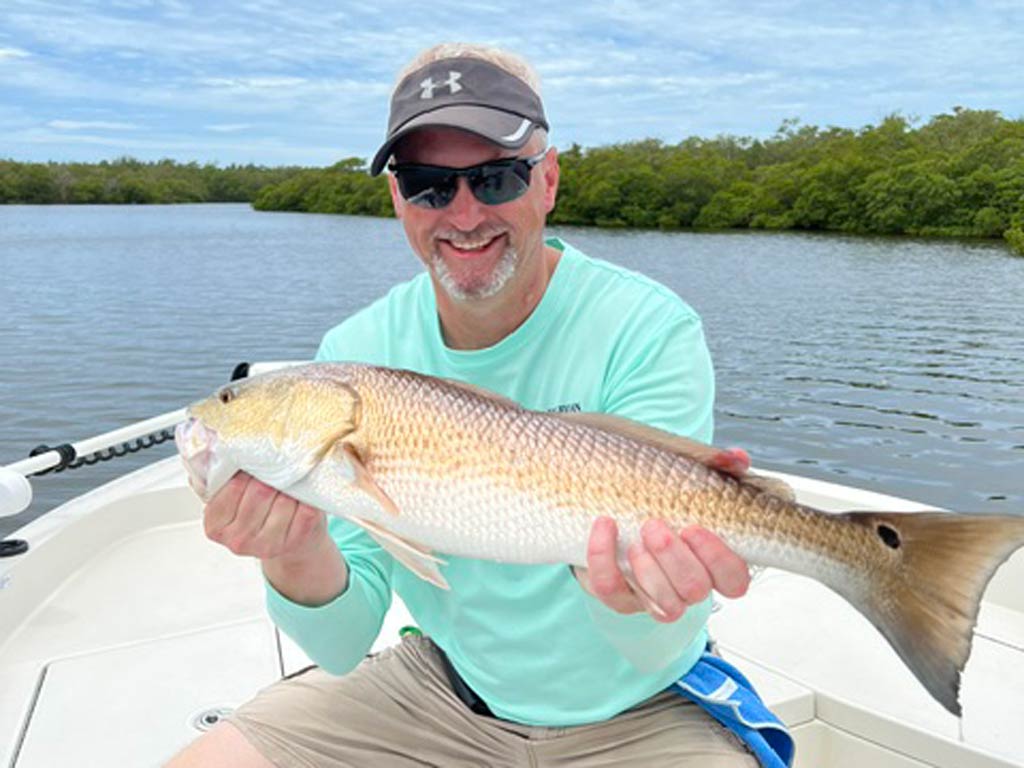 A man who's wearing sunglasses and a cap holds a large Redfish while sitting in a boat, with trees in the background.