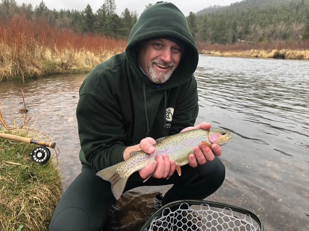 A man in a dark hoodie kneels by a river, smiling as he holds a freshly caught Trout with both hands. A fishing rod and net are nearby.