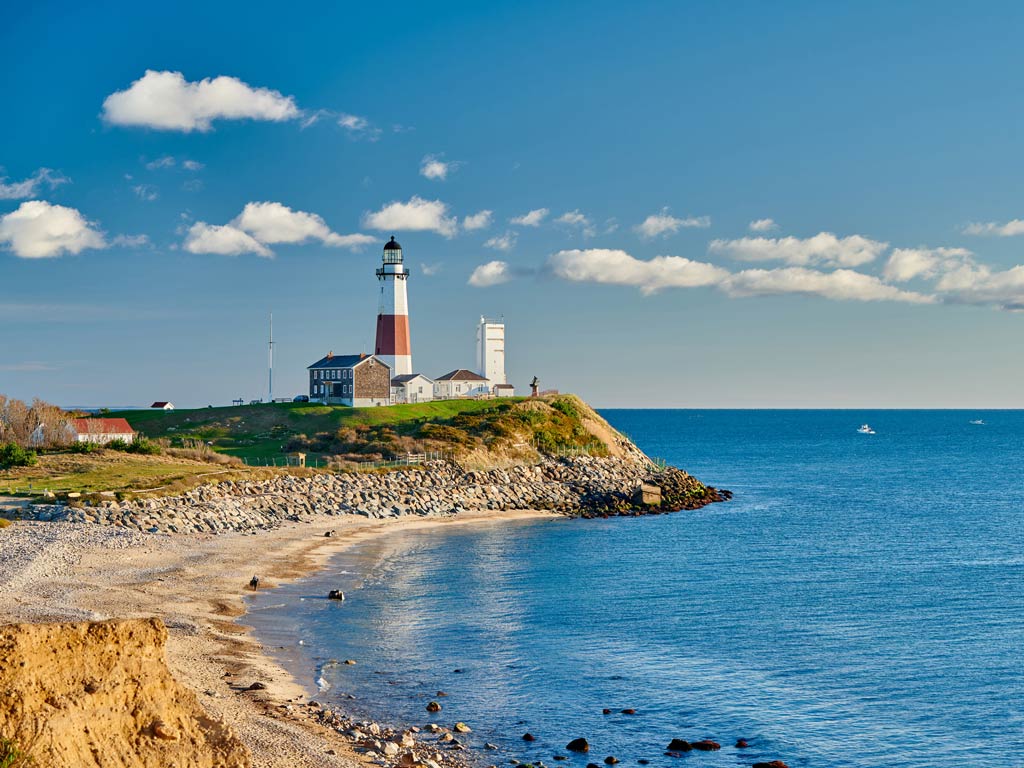 A lighthouse sits on a rocky cliff above a calm blue sea, it’s one of the most famous Montauk landmarks – Montauk Point Lighthouse