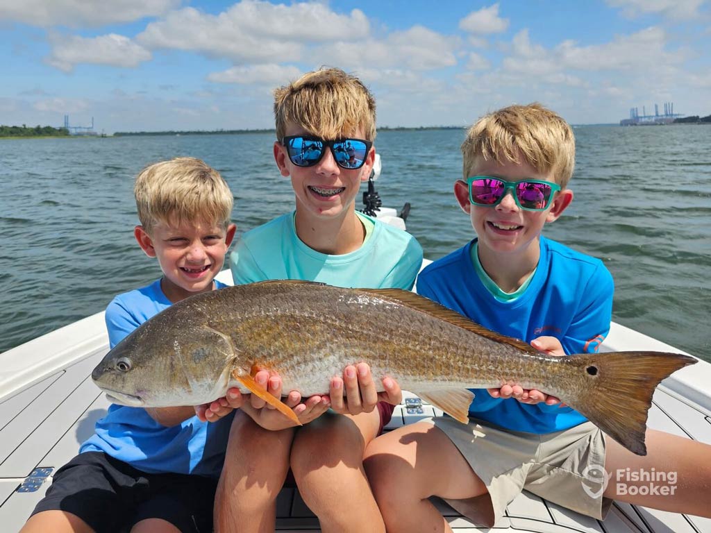 Three young boys sitting on a charter boat inshore and holding a large Redfish together, smiling at the camera during their Memorial Day fishing trip in one of the top spring destinations – Charleston