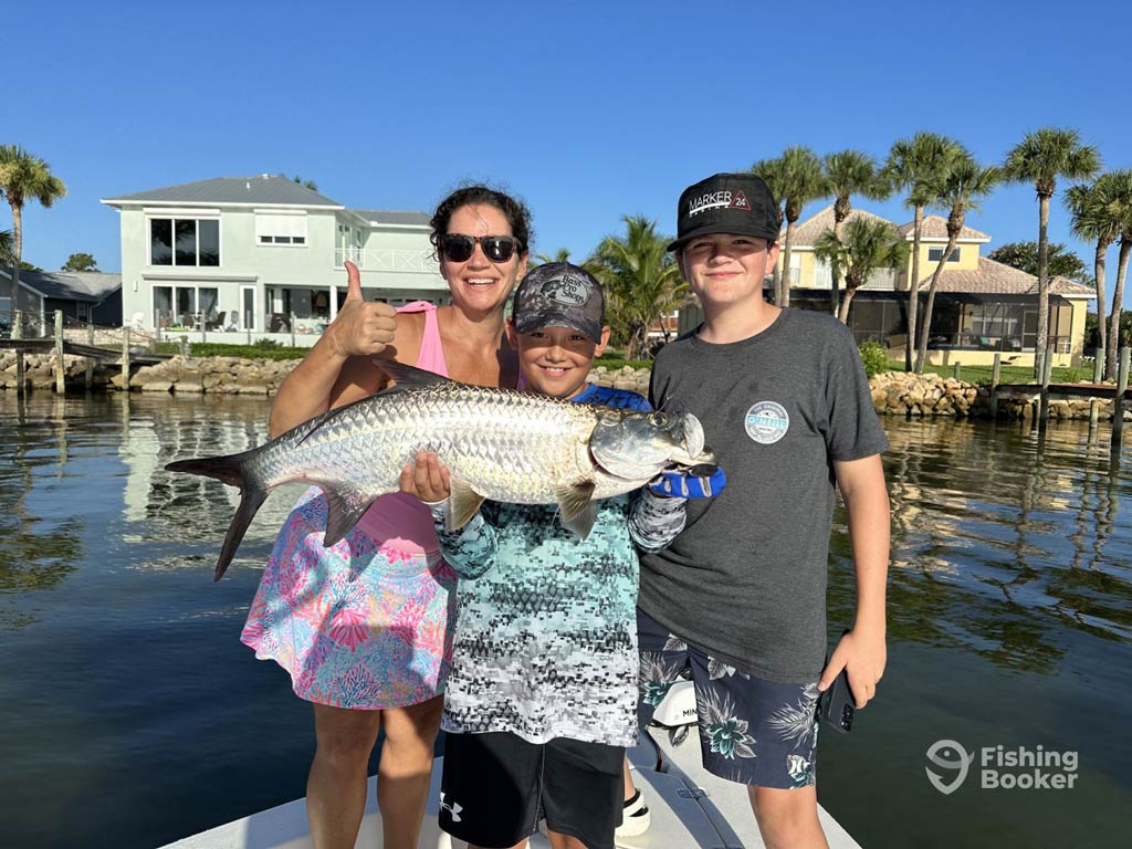 A family of three – a mom, a dad, and a kid – are standing on a charter boat and posing with a Tarpon they caught while fishing out of Orlando in Florida. They’re smiling while waterfront houses and palm trees are in the background.