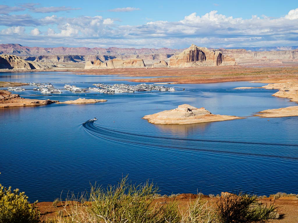 You’re seeing a wide view of Lake Powell, a top Memorial Day fishing destination, with a marina, boats, blue water, rocky islands, and desert cliffs under a partly cloudy sky.