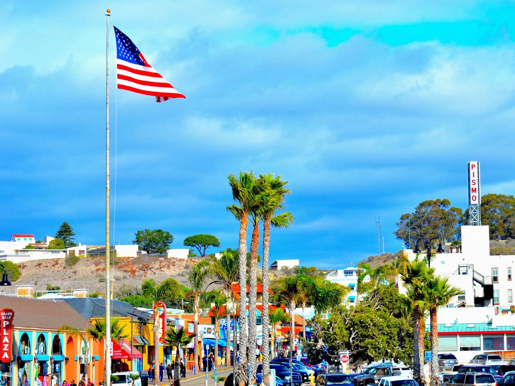 A tall American flag flies over a lively street lined with palm trees and shops in Pismo Beach, one of California’s top spring destinations.