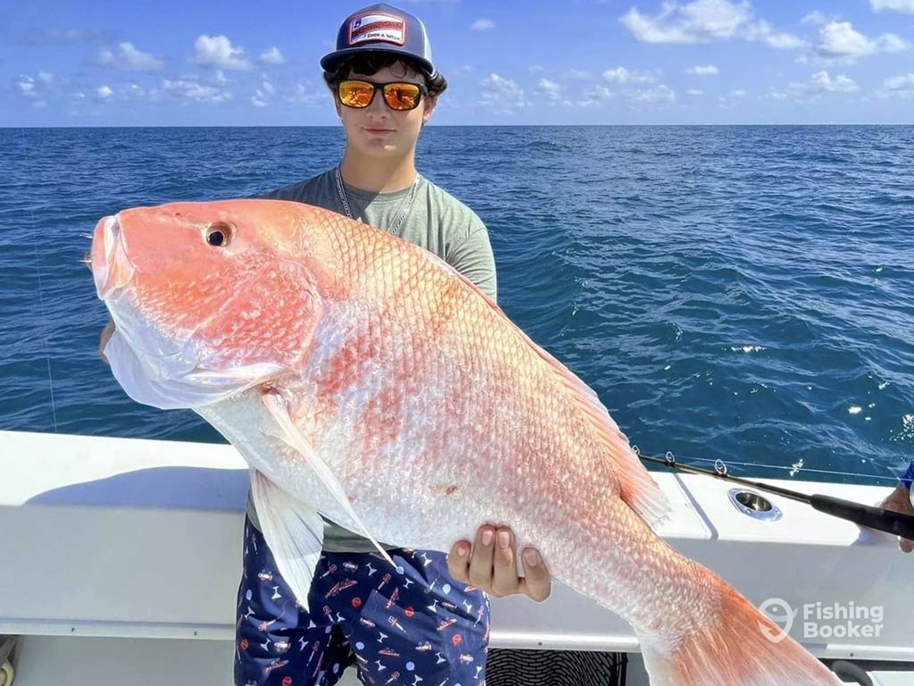 A young angler is standing on a charter boat and holding a huge Red Snapper, caught while bottom fishing offshore in Galveston, one of the top Red Snapper fishing destinations in the world.