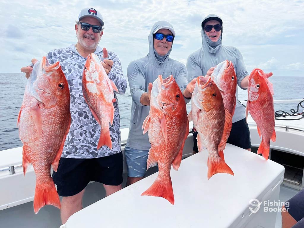 Three anglers are standing on a charter boat and showing off large Red Snappers they caught, smiling for the camera - a perfect day in Galveston, one of the top Red Snapper fishing destinations in the world. Ocean and cloudy sky in the background.