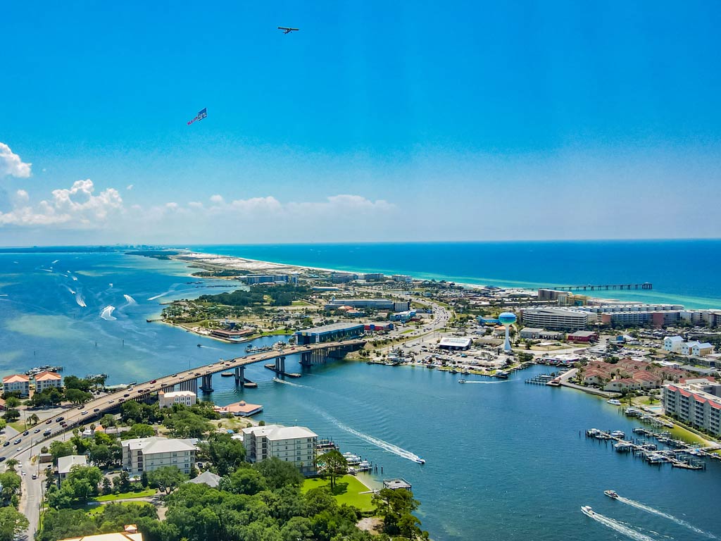 An aerial view of the coastal city of Fort Walton Beach known for its Red Snapper offer, with boats in the bay, a bridge, shoreline buildings, and planes soaring across a clear blue sky.
