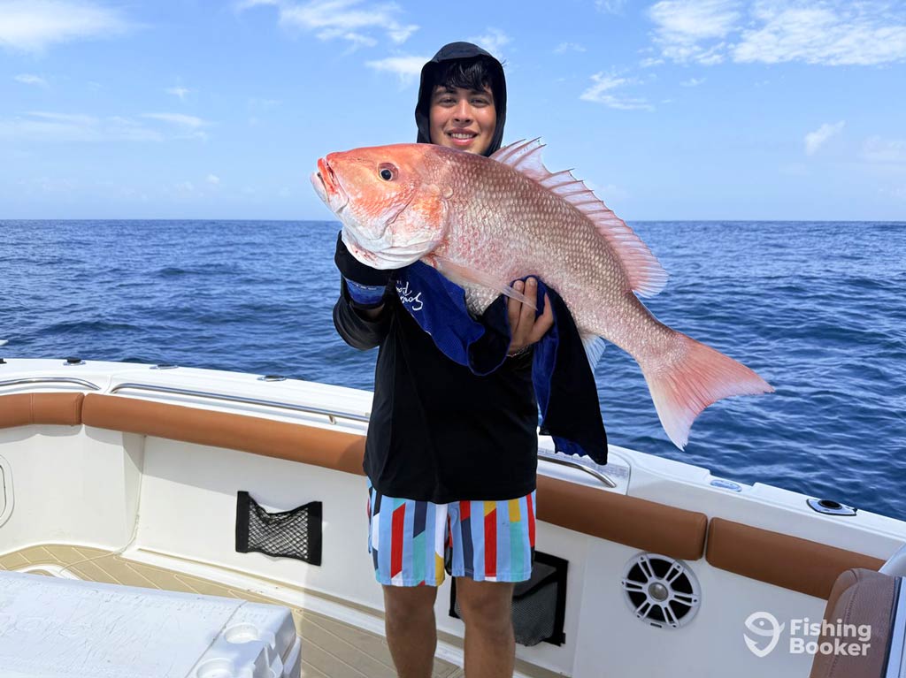 A teenage angler is standing on a charter boat holding a large Red Snapper, showing off his bottom fishing catch from the Gulf during the high Red Snapper season.