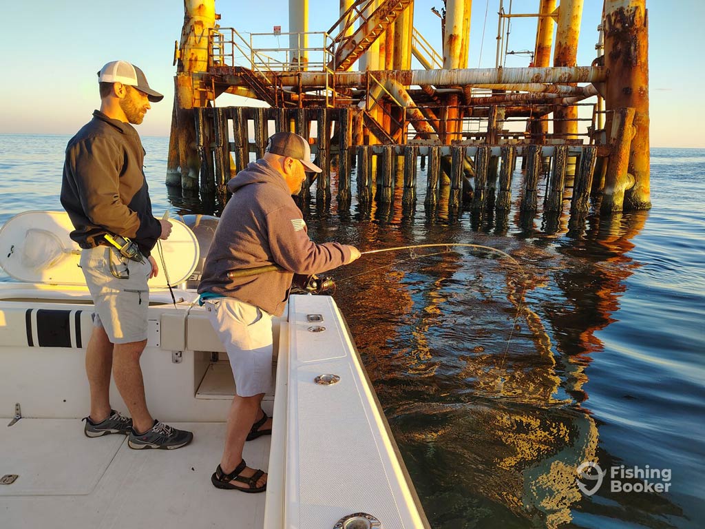 Two anglers are bottom fishing from a charter boat near a rusty offshore platform resembling a smaller oil rig, with one reeling in a catch.