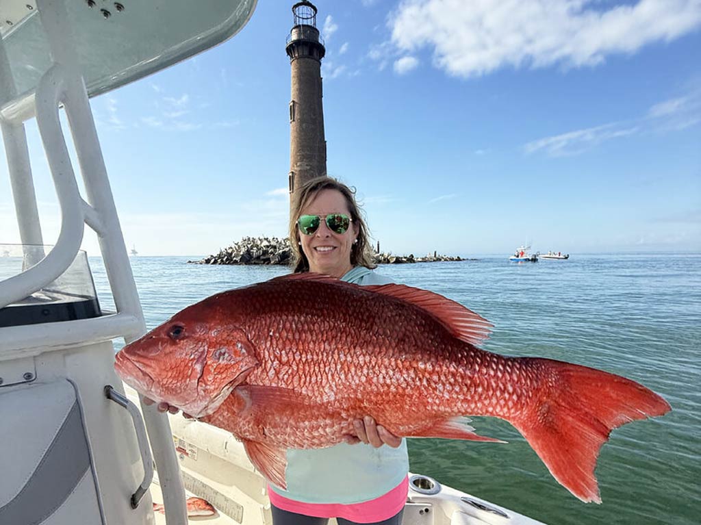 A female angler standing on a charter boat and holding a large Red Snapper, showing off her Dauphin Island catch, with a lighthouse and rocky shore in the background.
