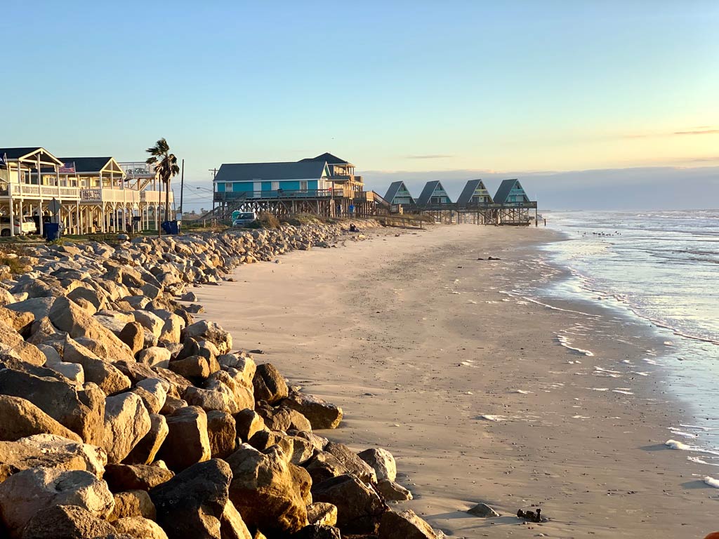 A view of Surfside Beach with the rocky shoreline and sandy beach with stilt houses along the water’s edge, it’s an ideal sunrise spot, often sought after in Freeport.