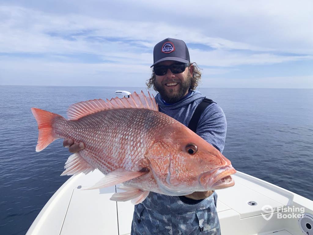 An angler is wearing sunglasses and a cap as he holds a large Red Snapper while posing on a charter boat out in the open water during the Atlantic Red Snapper season, with a cloudy sky overhead.