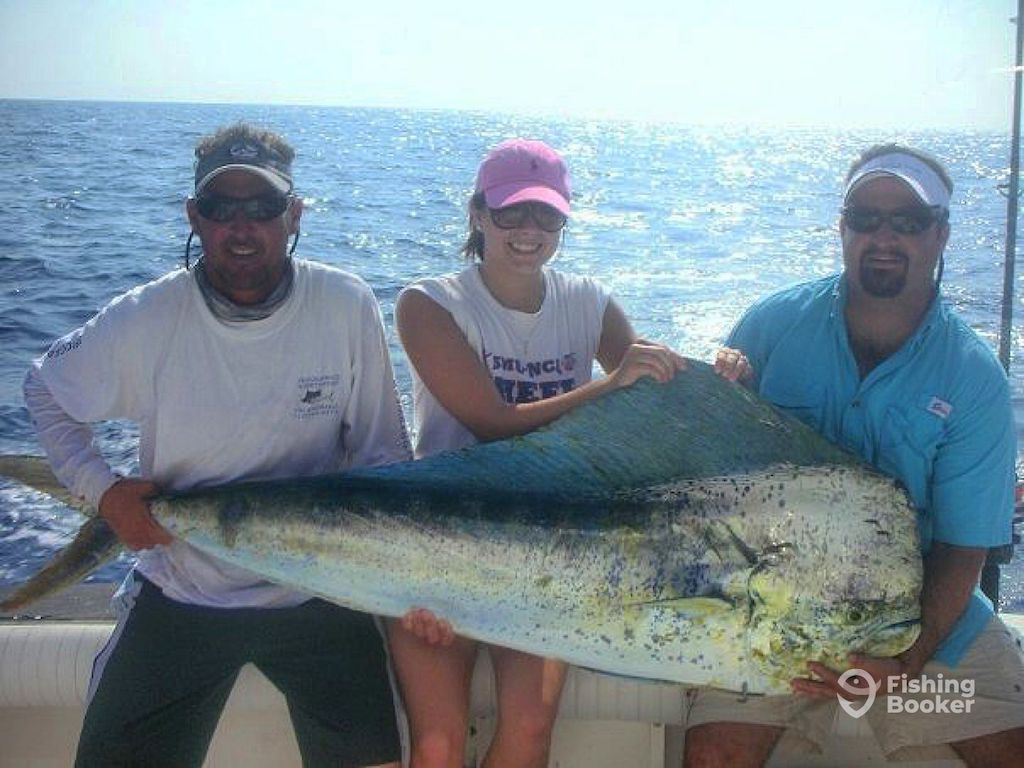 Three people on a boat hold a large Mahi-Mahi with the ocean behind them, showing off an epic catch during Florida Mahi Mahi Season. "Fishing Booker" logo's at the bottom right.
