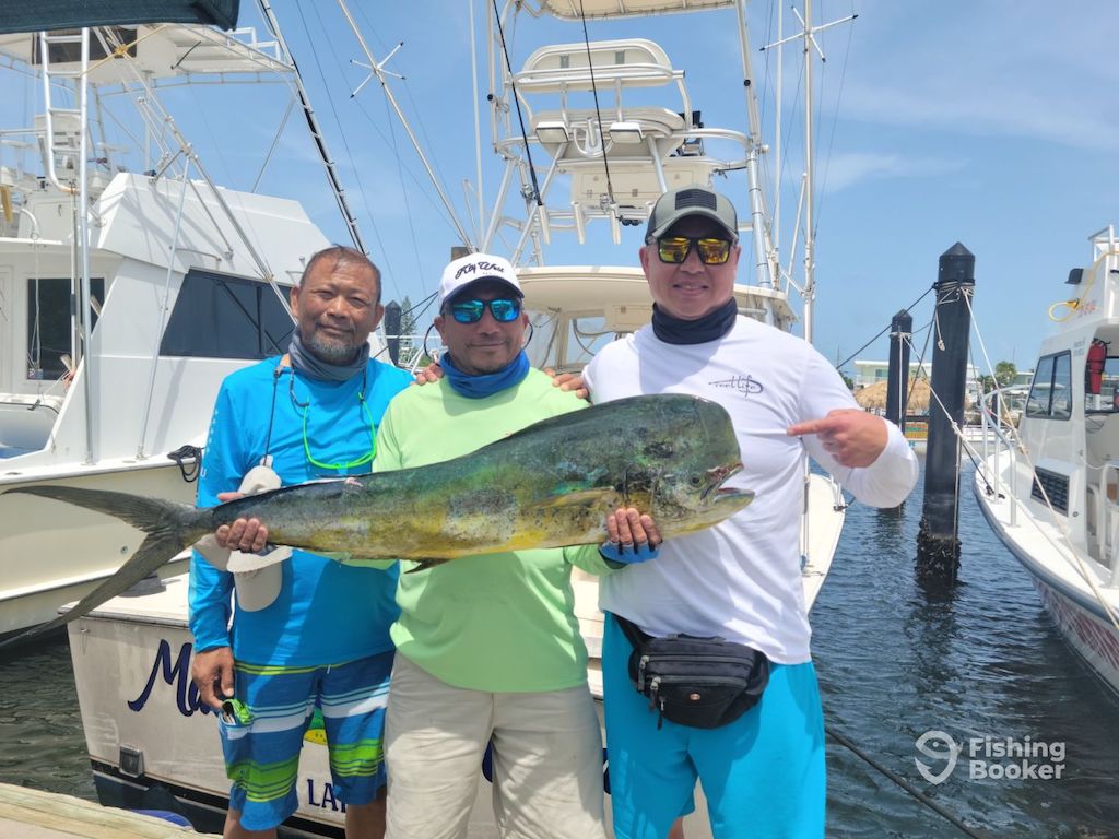 Three men pose on a marina dock, holding a large Mahi Mahi caught during Florida season. Boats are in the background, and they're wearing sun-protective clothing and hats.