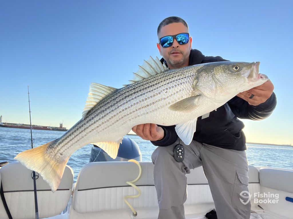 A man holding a big Striped Bass on a boat during the season, wearing sunglasses and outdoor clothes, with water and sky behind him.