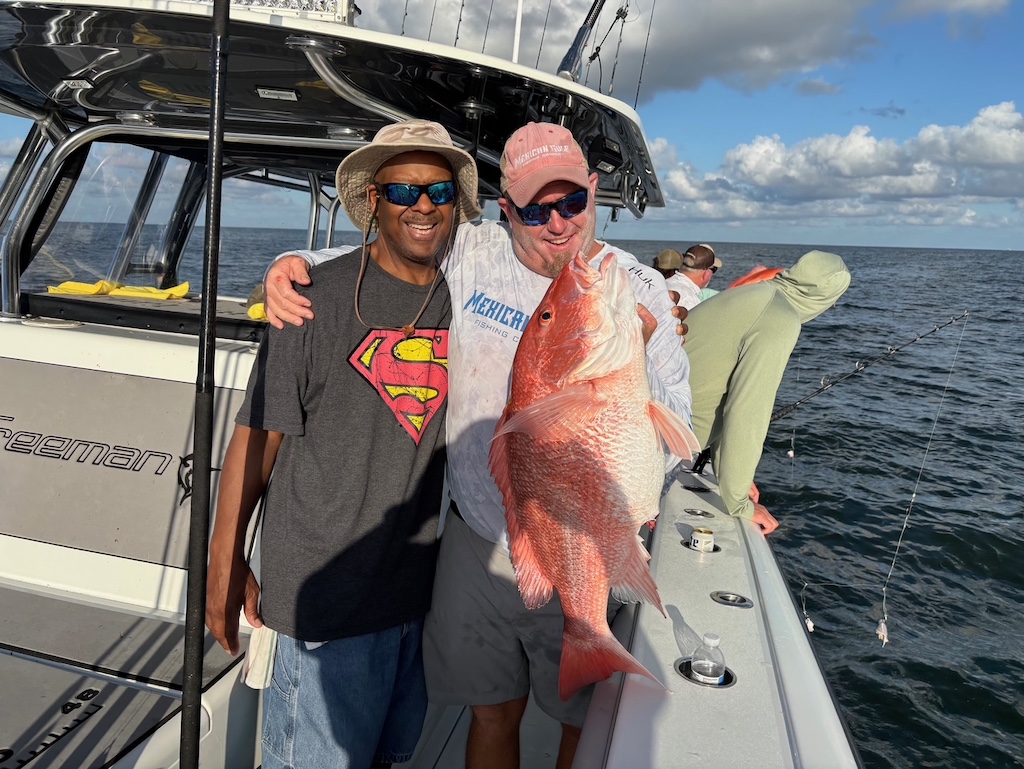 Two men are on a boat; one is holding a large Red Snpper, and they’re both smiling at the camera. Offshore fishing action goes on in the background on a sunny day.