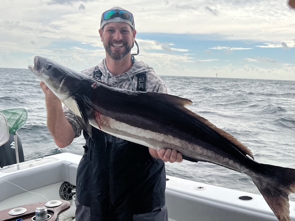 A man standing on a boat, holding a large Cobia with dark and light stripes, showing off offshore fishing action against the ocean and a cloudy sky.