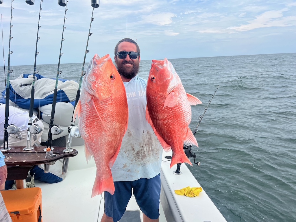 A man is standing on a boat, holding two large Red Snapper fish, showing off some offshore fishing action with rods and the ocean in the background.