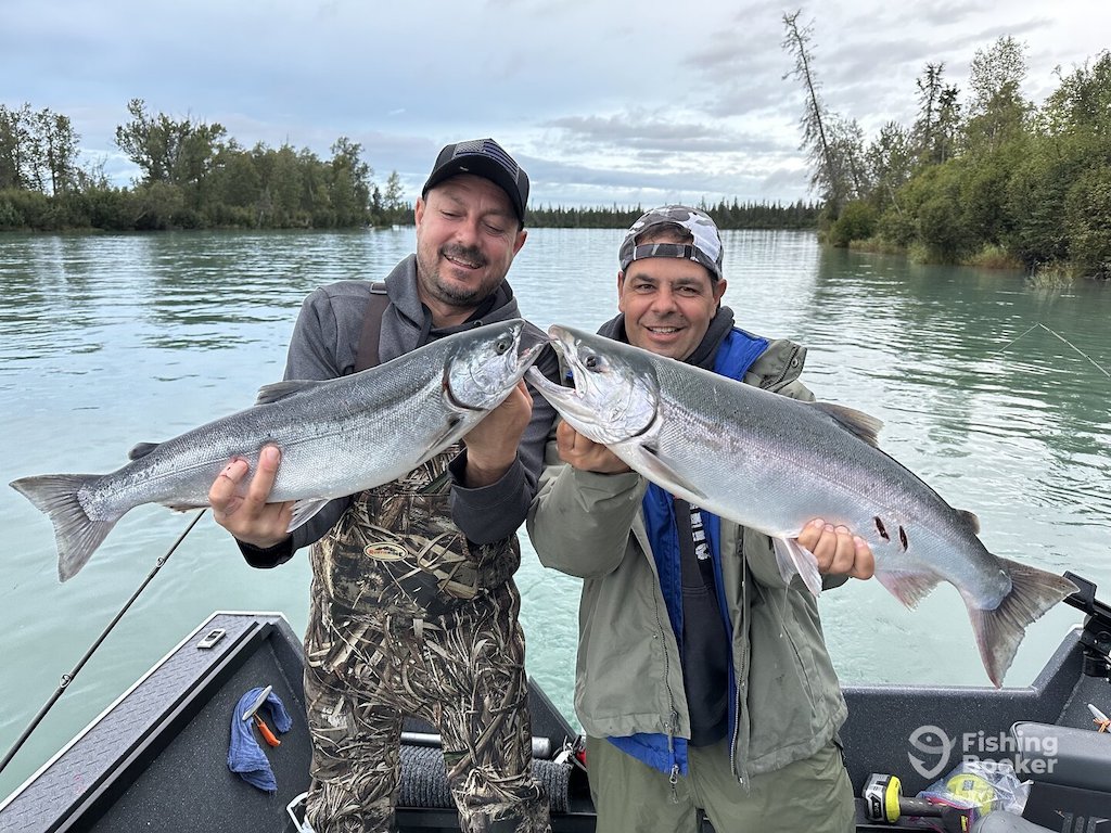 Two men stand on a boat holding large Salmon, facing each other and celebrating a successful Alaska Salmon season. There's a lake and trees in the background.