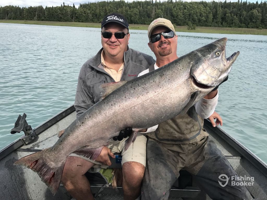 Two men sit in a boat on a lake during Alaska Salmon season, with one proudly holding a large Salmon; trees and water fill the scenic background.