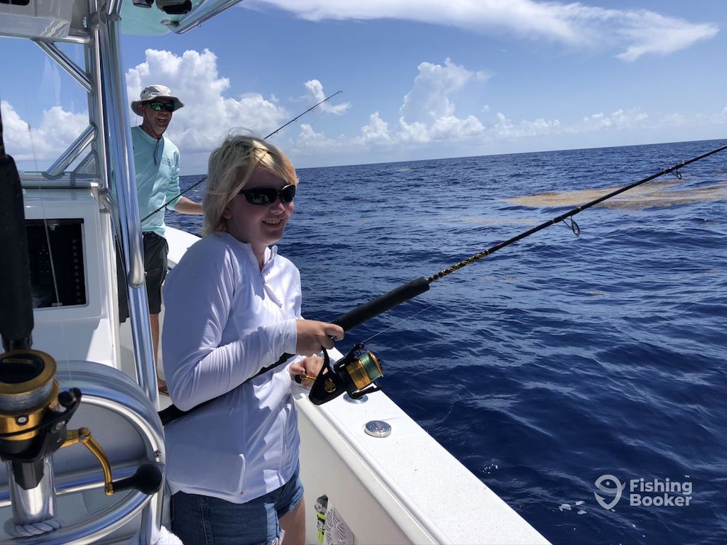Two people are fishing from a boat in the ocean under a partly cloudy sky. One is holding a fishing rod while the other's nearby, both wearing sunglasses and casual clothes.