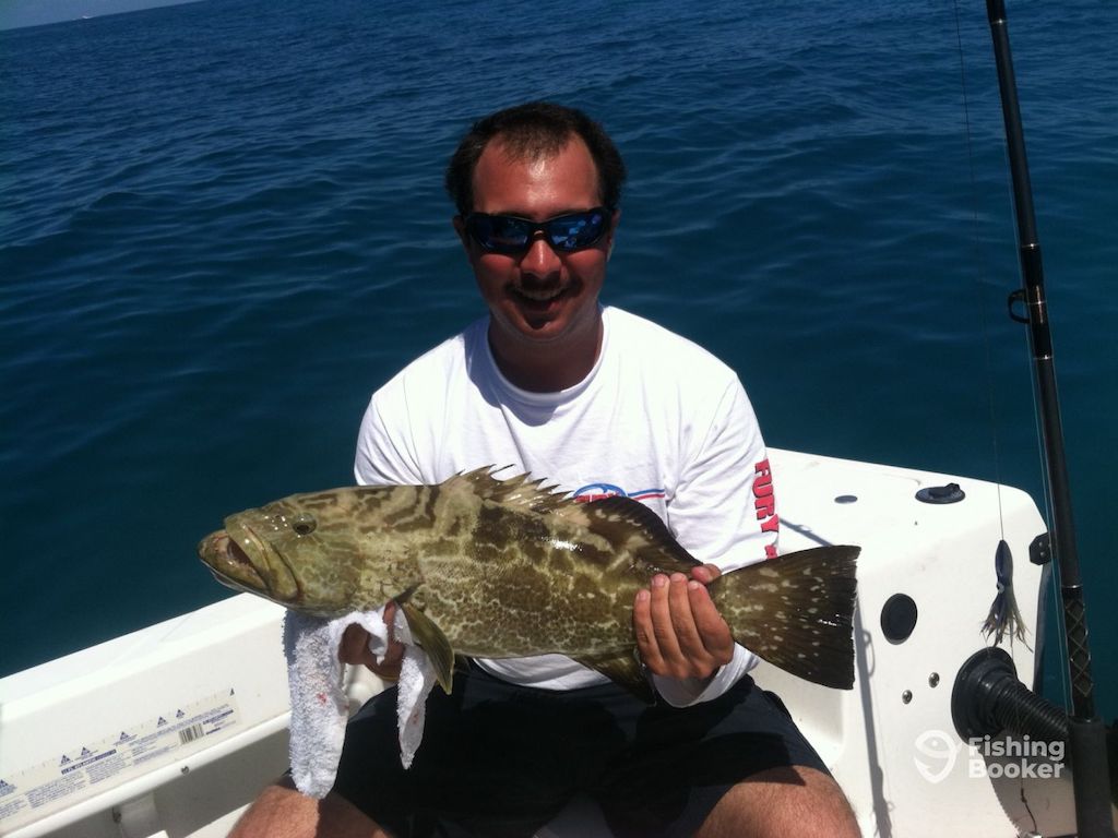 A man sitting on a boat, holding a Gag Grouper, with the ocean behind him.
