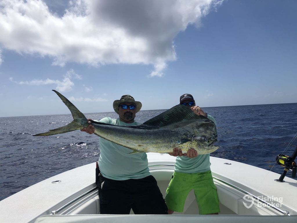 Two people on a boat holding a large Mahi Mahi, with the ocean and sky in the background.