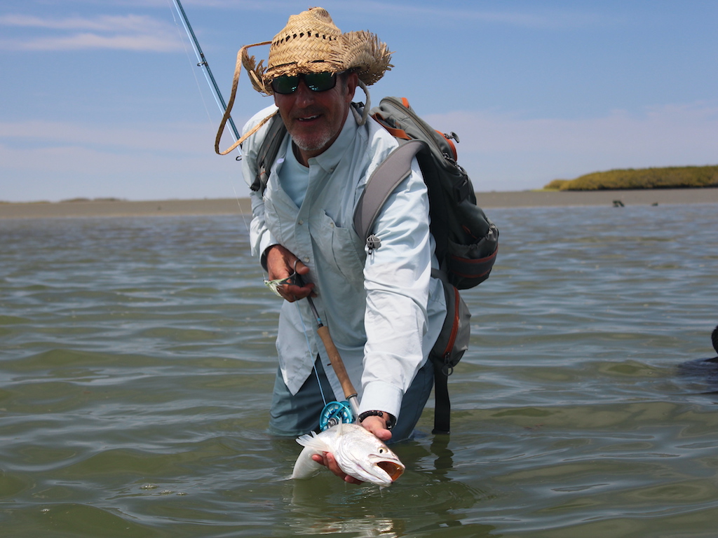 A man in a straw hat and sunglasses wade fishing and showing off his catch to the camera on a clear day.