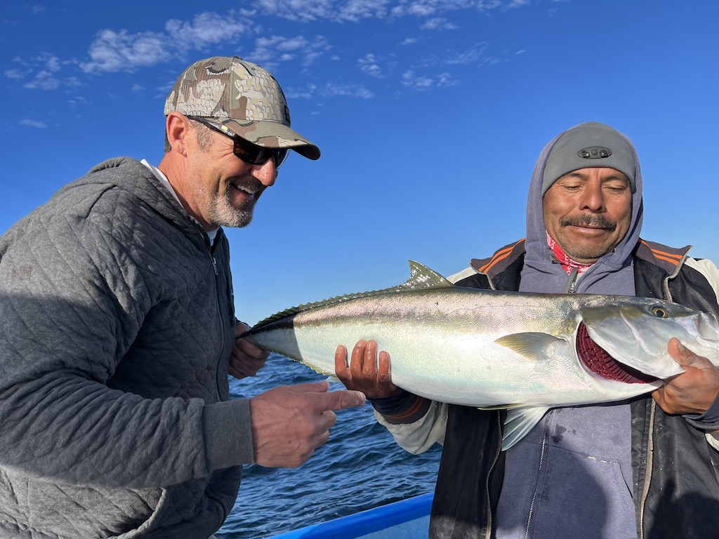 Two men smile as they struggle to hold a Yellowtail after a successful fishing trip near Loreto.
