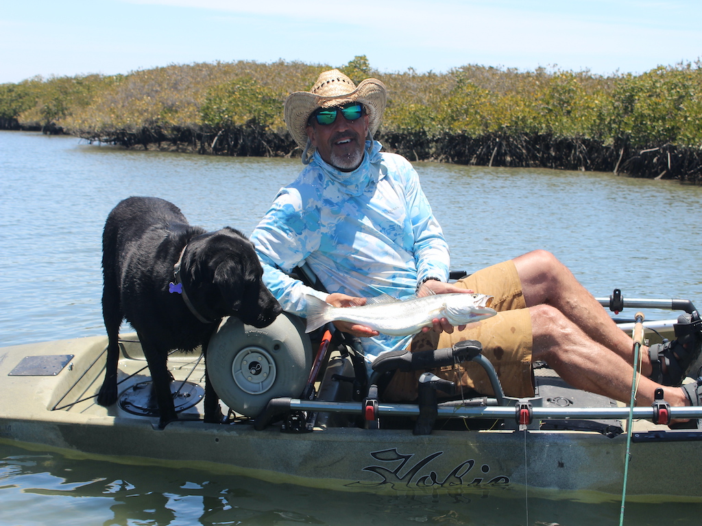 A view across the water towards a man and a black labrador on a kayak, as the man shows off a fish he caught.