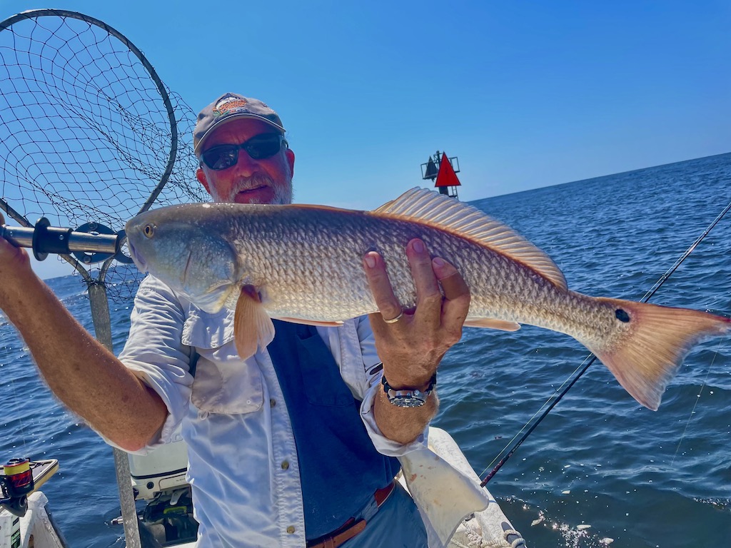 A man on a boat holding a Redfish in front of his face on a clear day with the water visible behind him.