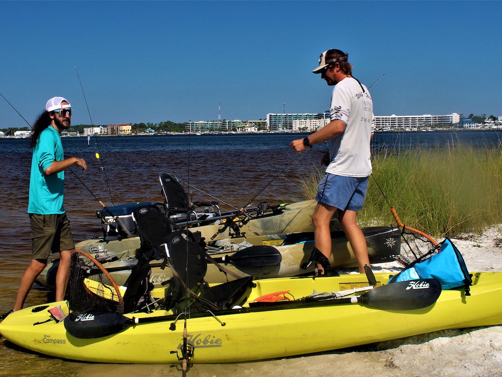 Two men standing on kayaks on shore in Orange Beach, AL, ready to go fishing the shallows.
