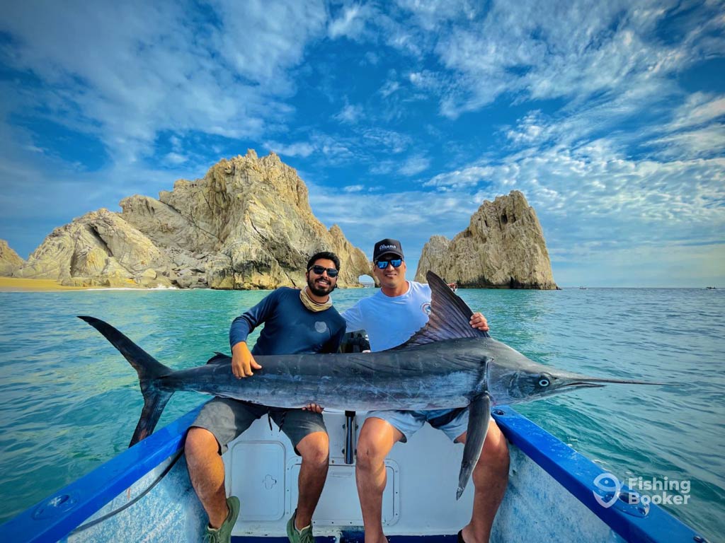 Two anglers are sitting on a charter boat and holding a large Marlin against rocky cliffs and clear blue water, sharing funny fishing saying under a partly cloudy sky.
