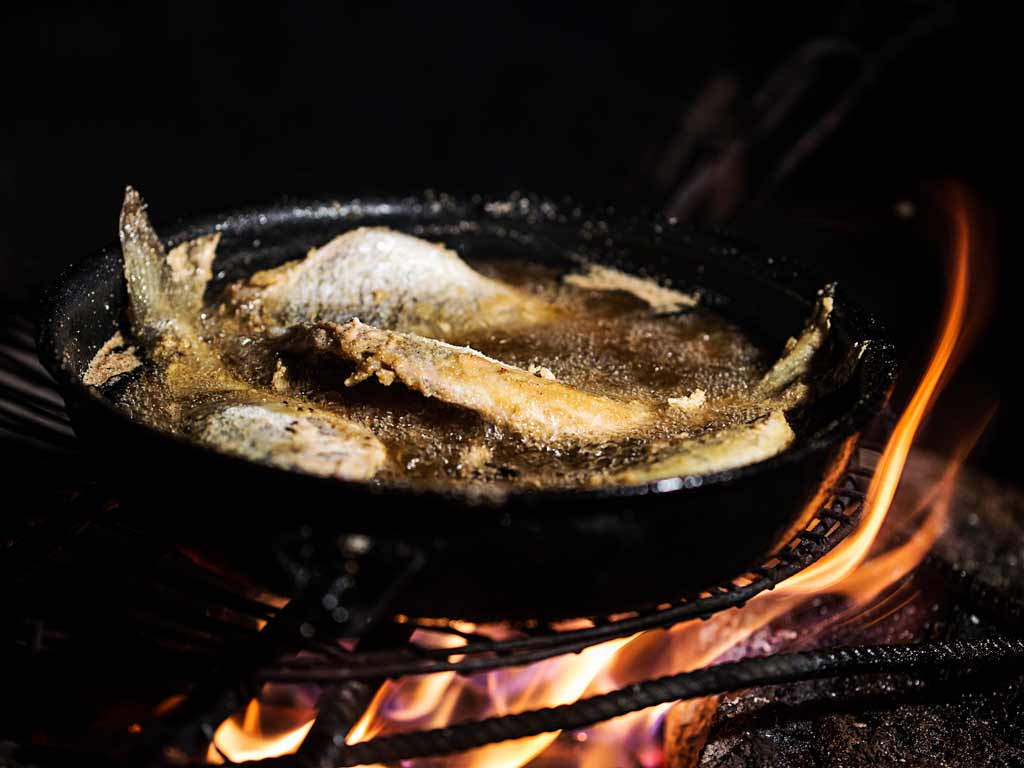 Fish are frying in oil in a cast iron pan over an open flame, with visible fire and grill underneath.