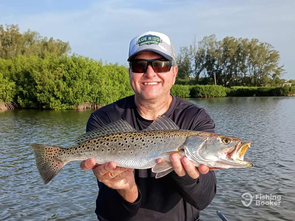 An angler is wearing sunglasses and a cap, while standing on a boat in calm water lined with trees and posing with a Speckled Trout he caught in Florida.
