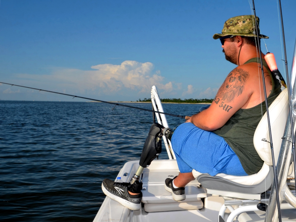 A man with a prosthetic leg sitting on a boat fishing in open water under a clear sky, wearing a hat, sunglasses, and casual clothes.