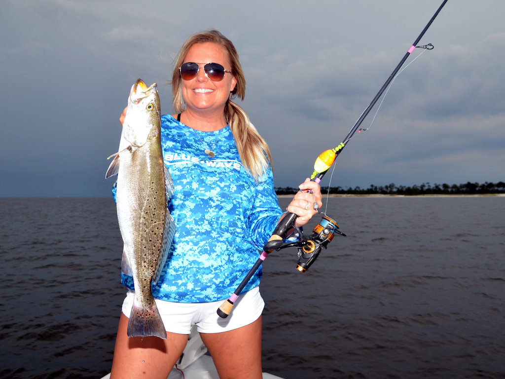 A woman in sunglasses and a blue patterned shirt standing on a boat, holding a fishing rod and a large Speckled Trout, with water and a cloudy sky in the background.