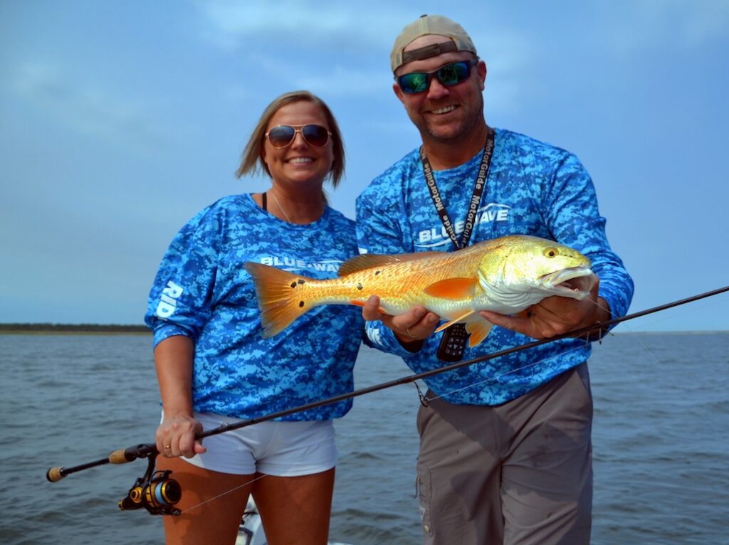 Two people in matching blue shirts are on a boat, holding a fishing rod and showing off a large, freshly caught Redfish over open water.