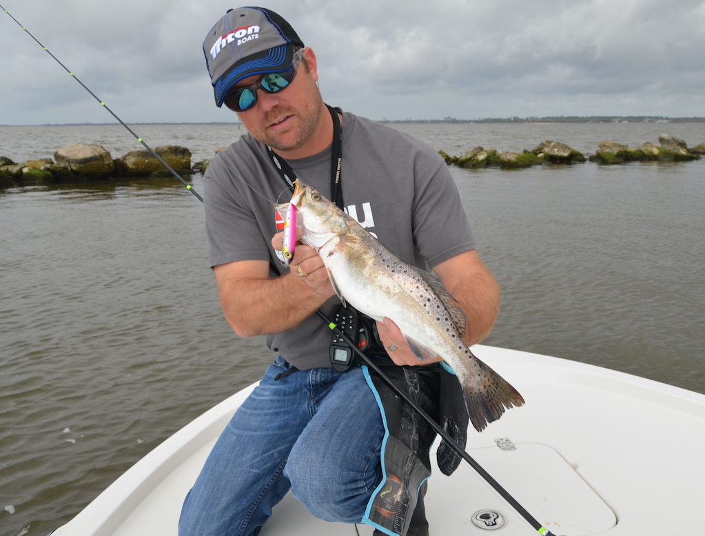 A man kneeling on a boat, holding a freshly caught Speckled Trout along with fishing gear. Water and rocks are in the background.