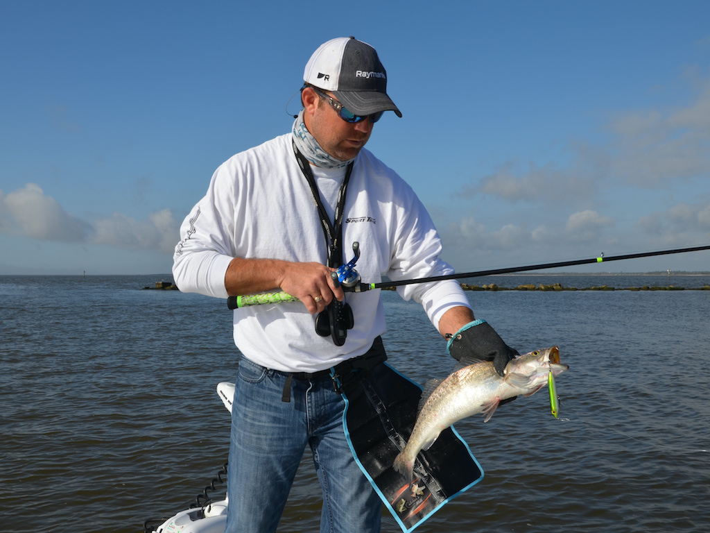 A man standing in shallow water, holding a fishing rod in one hand and a Speckled Trout in the other, with a lure still in the fish's mouth.