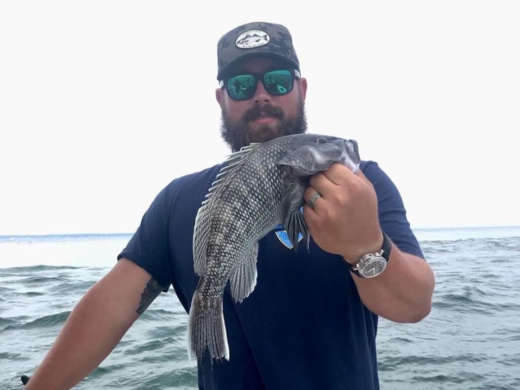 A man wearing sunglasses and a cap holds a Black Seabass in one hand while standing on a boat with the ocean in the background.