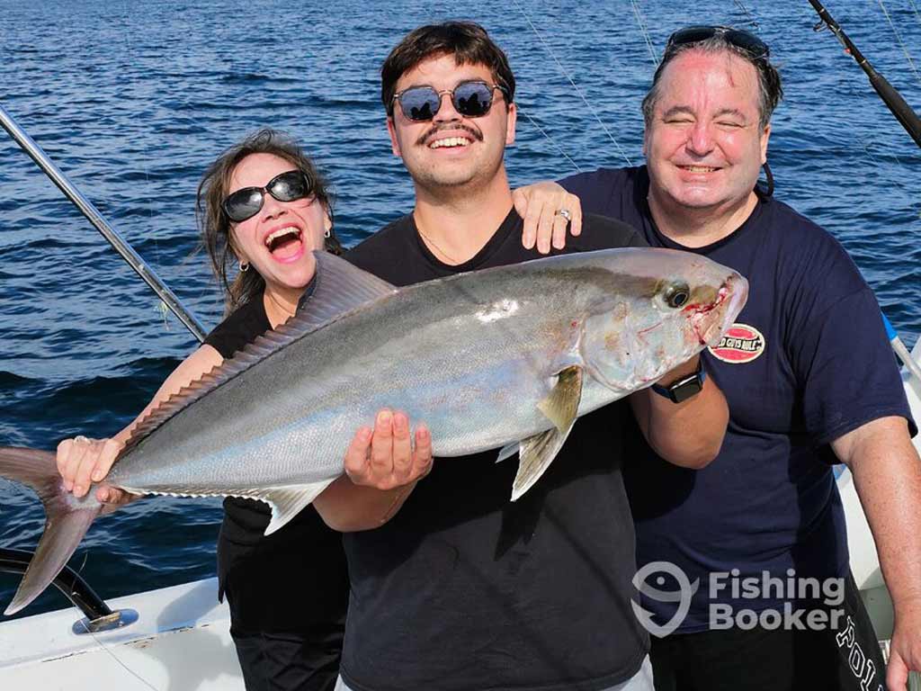 Three anglers are standing on a charter boat and holding a large Greater Amberjack while smiling at the camera with the ocean behind them