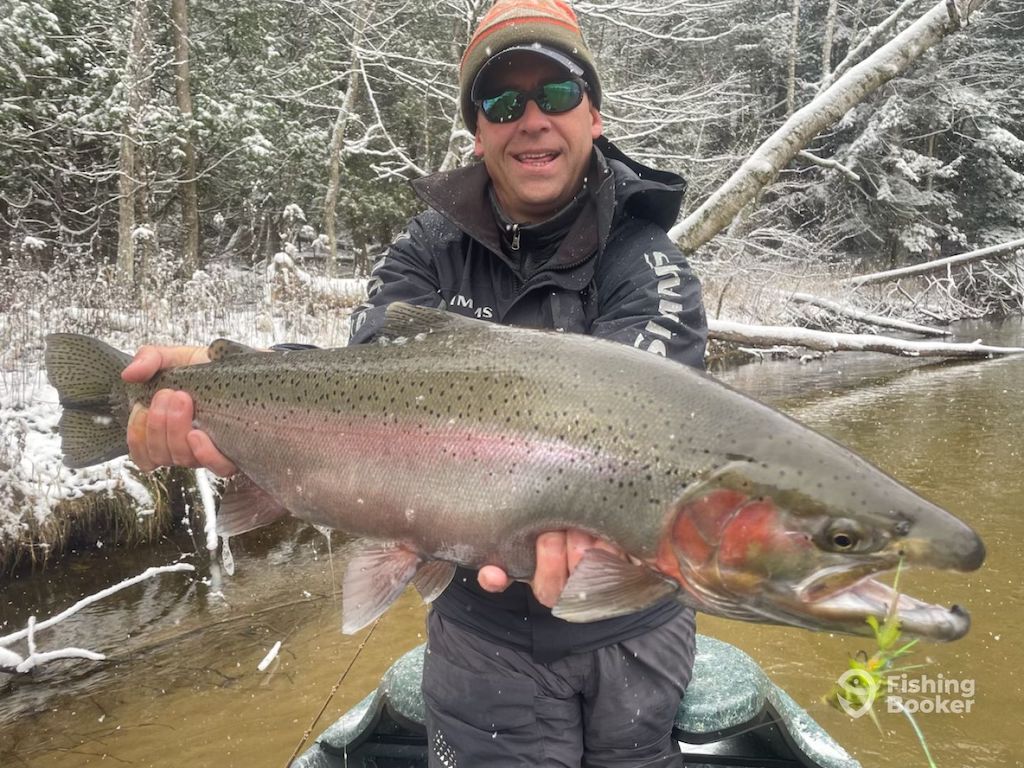 A man wearing sunglasses and winter clothes while holding a big Rainbow Trout they caught during Michigan Trout season by a snowy riverbank in a forested area.