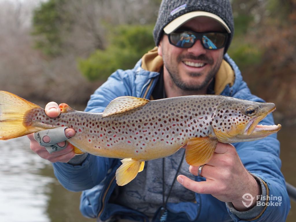 A man in outdoor clothing holding a Brown Trout with both hands, smiling at the camera by a body of water during Michigan Trout season.