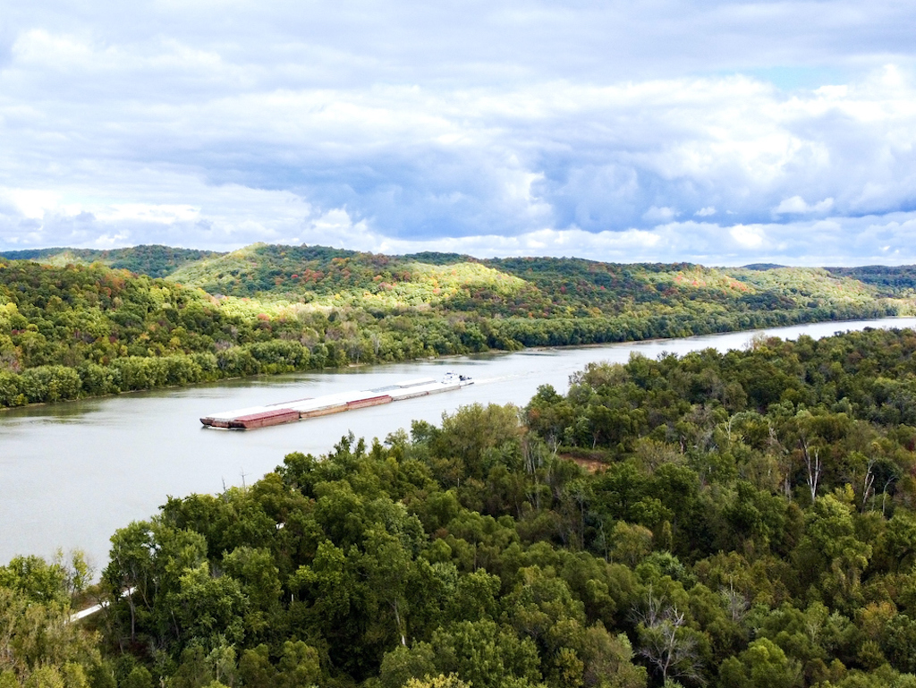 A barge moving along a wide river, surrounded by dense, green forest and rolling hills under a partly cloudy sky during Michigan Trout season.