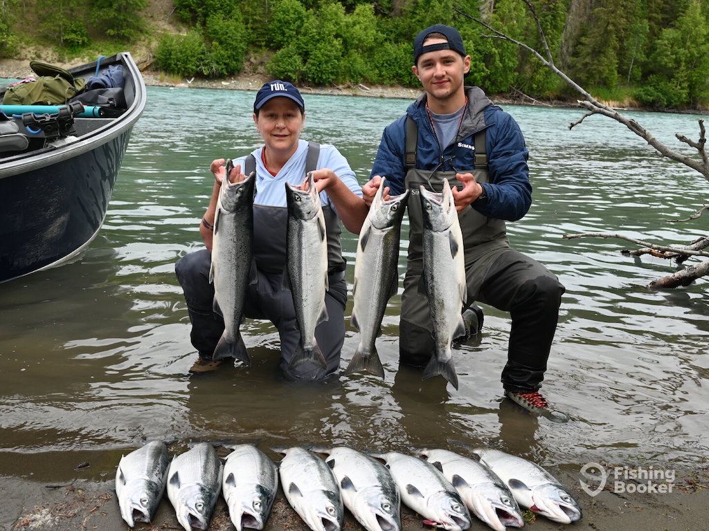 Two people kneel by a river during Washington Salmon season, each holding four large Salmon, with eight more laid out nearby.