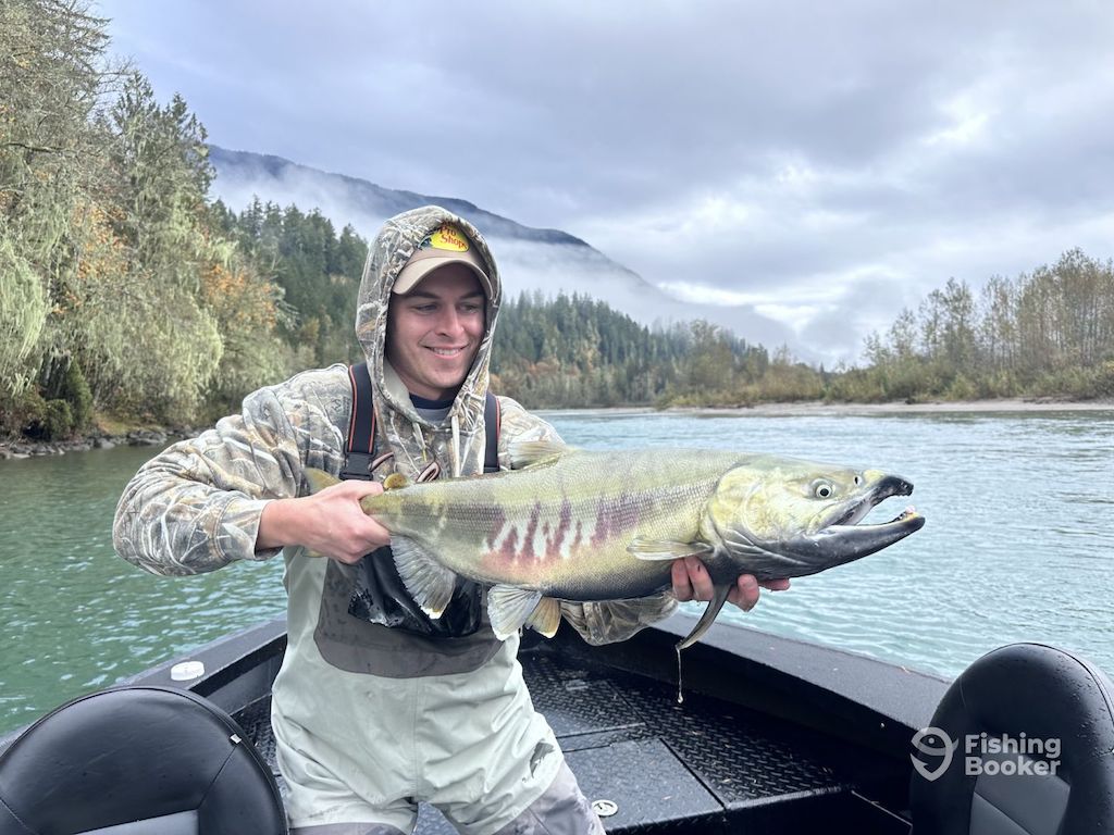A person in camo clothing stands on a boat holding a large Salmon, showing off Washington Salmon season's beauty with a river, forest, and mountains behind. The fishing Booker logo is in bottom right.