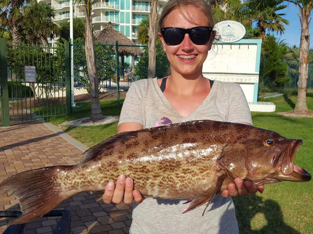 Angler is wearing sunglasses and holding a large Scamp Grouper outdoors by a fence and palm trees on a sunny day after fishing in the South Atlantic.