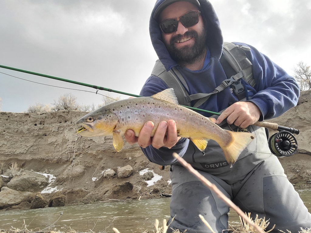 A man in fishing gear holding a Brown Trout near a riverbank, with a fishing rod in one hand and a cloudy sky in the background.